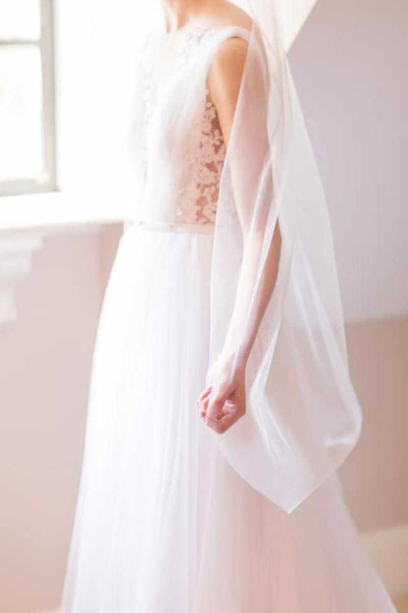 Bride in white lace-bodice gown holding sheer veil, soft natural light from window