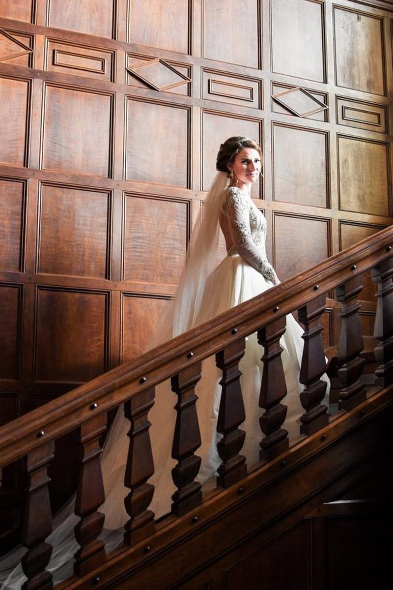 Bride with veil ascending grand wooden-paneled staircase inside mansion