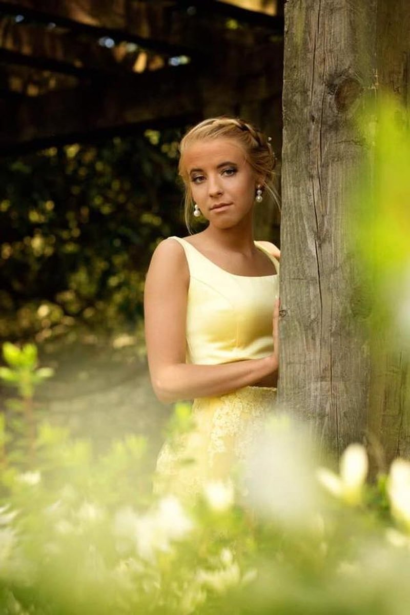Young woman in yellow dress leaning against wooden post amid garden greenery