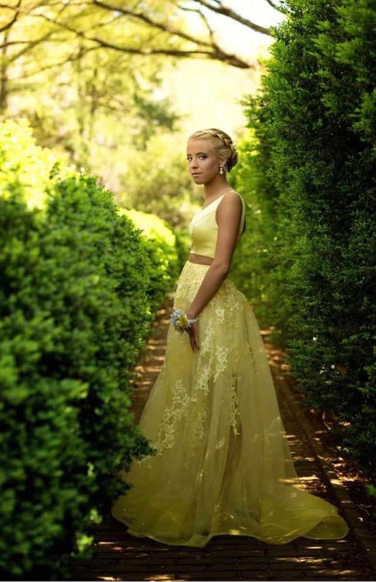 Young woman in yellow two-piece lace gown standing in manicured hedge-lined garden path