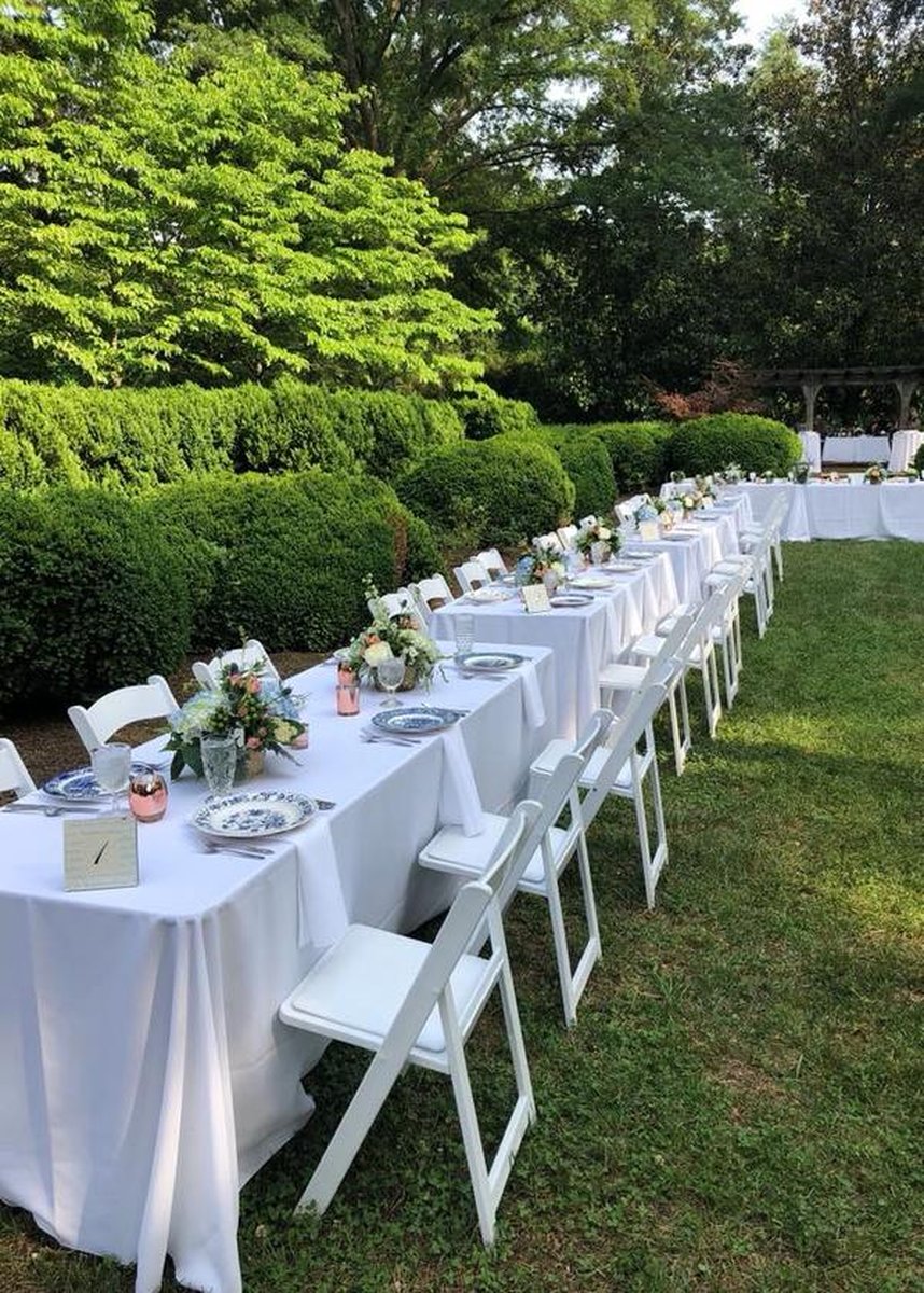 Long banquet table with white linens and folding chairs on lawn alongside boxwood hedges