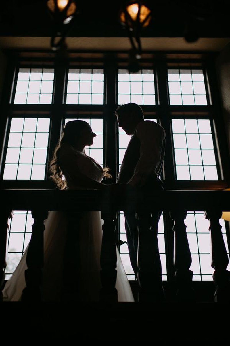 Silhouette of bride and groom on balcony with large mullioned windows backlit