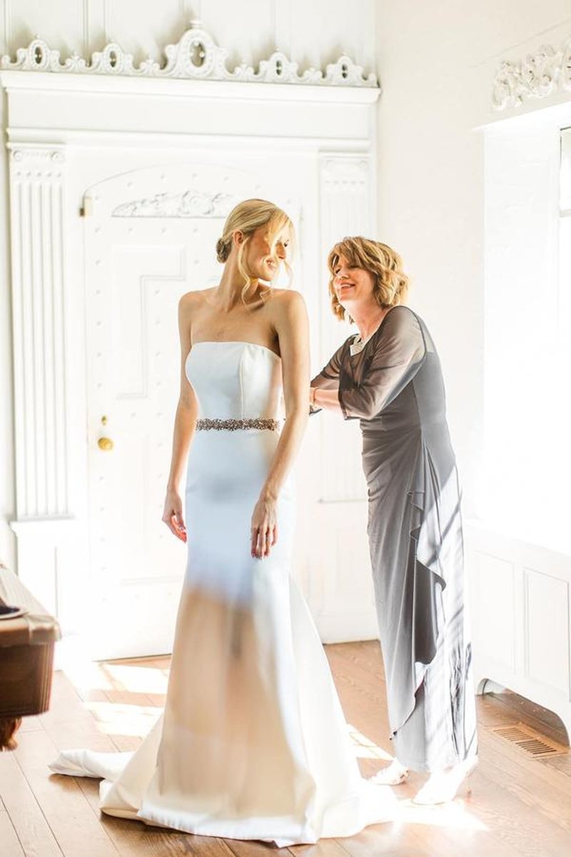 Mother helping bride into wedding dress in elegant mansion room with ornate white molding