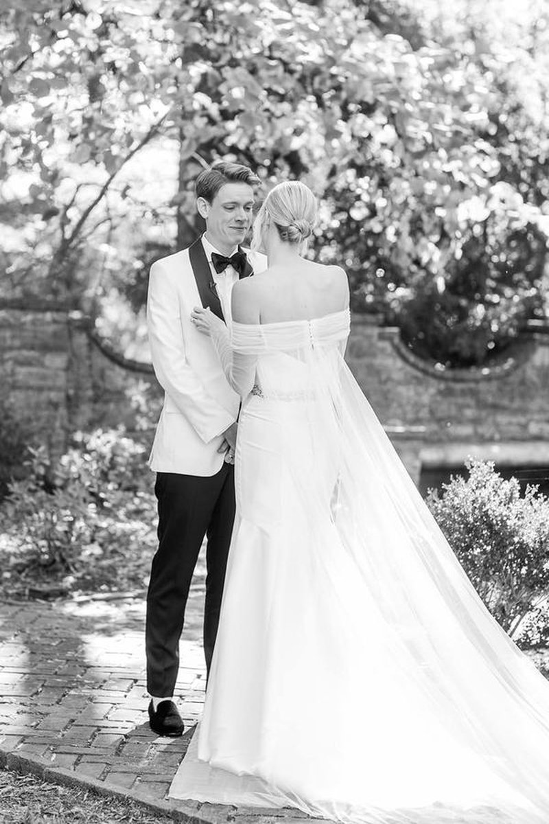 Black and white portrait of bride and groom on brick garden path, cape and train flowing