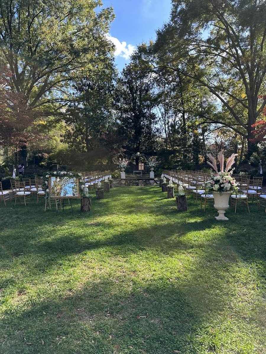 Outdoor wedding ceremony setup with gold chiavari chairs, floral urns, pampas grass, rustic log aisle markers