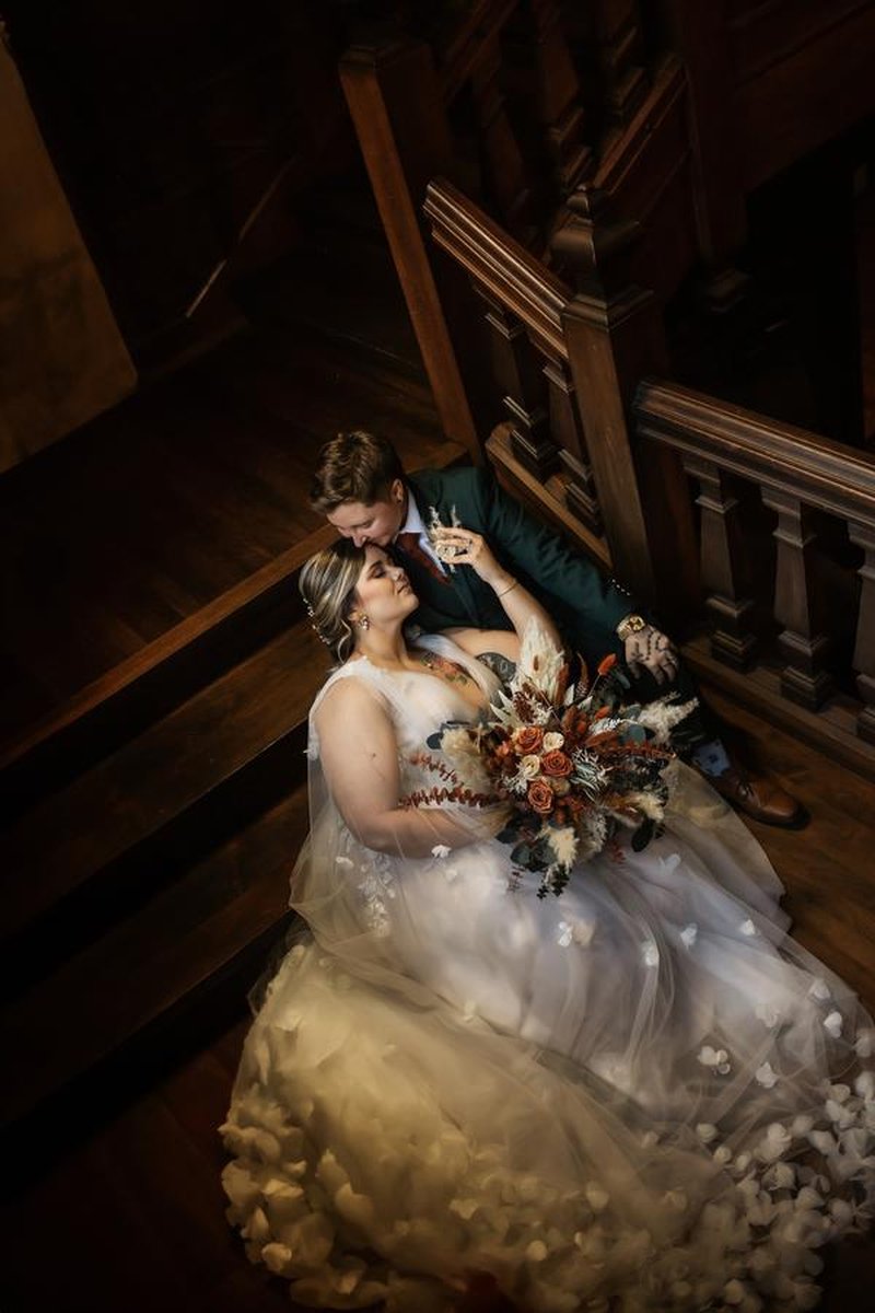Overhead shot of couple seated on grand wooden staircase, bride's tulle gown cascading down steps