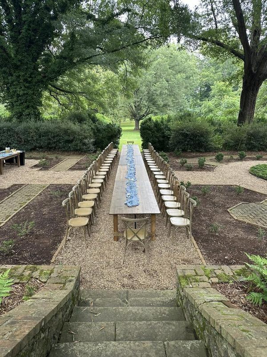 Long farmhouse dining table set in formal garden courtyard with cross-back chairs and blue runner