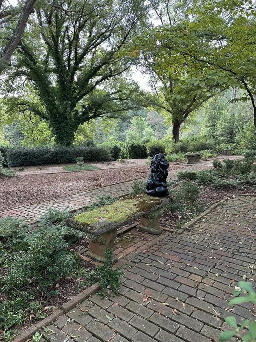 Brick pathway through lush garden with moss-covered stone bench and black lion statue