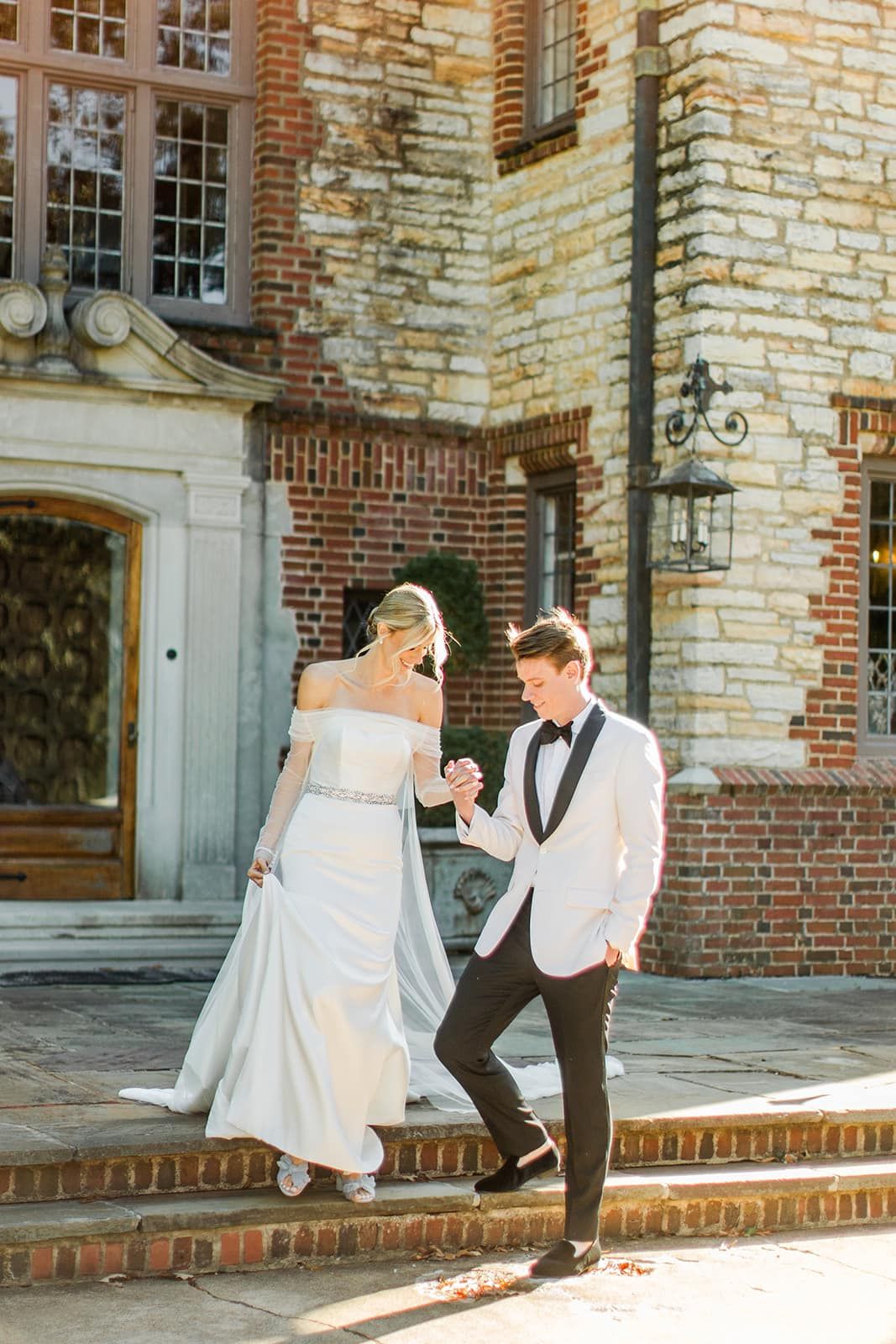 Bride and groom walking hand-in-hand down brick steps of stone mansion in golden hour