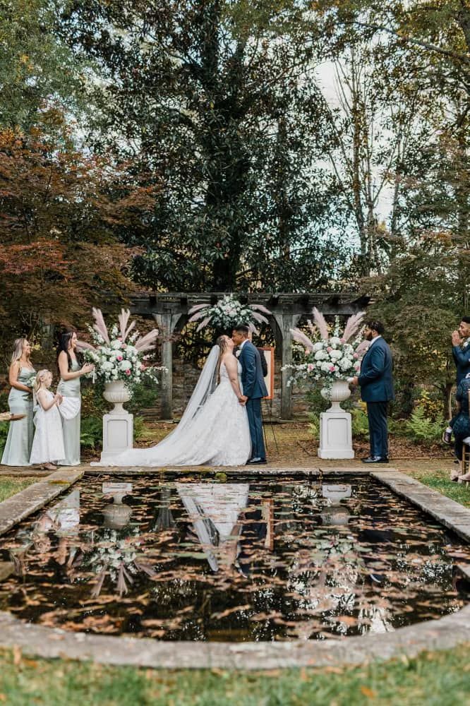 Bride and groom kissing by reflecting pool with stone pergola and pampas grass in autumn
